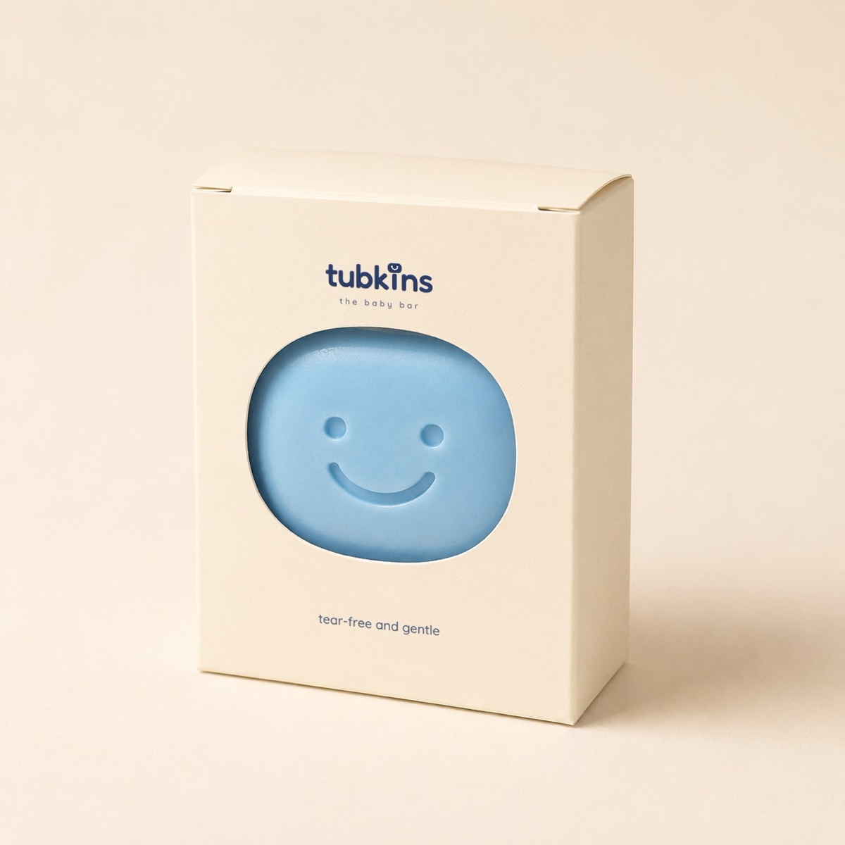 A cream, plastic-free Tubkins carton with a die-cut window revealing the smiling blue bar, the tubkins wordmark printed above and 'tear-free and gentle' below.